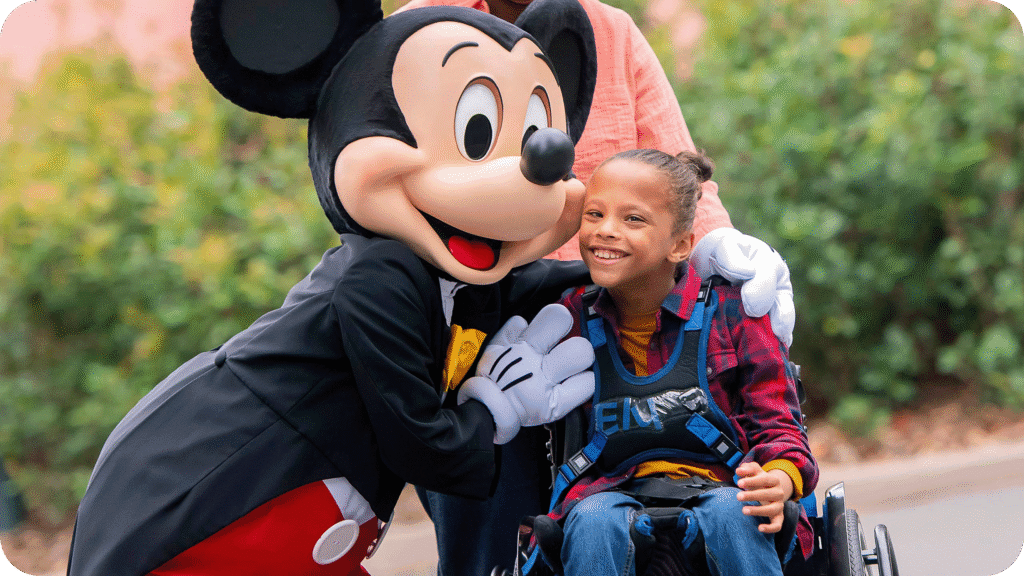 Une jeune fille en fauteuil roulant est accompagnée d'une personne vêtue d'une souris, soulignant ainsi l'esprit d'inclusion de Disneyland Paris et le soutien précieux apporté par un Accompagnant Handicap Disneyland Paris.