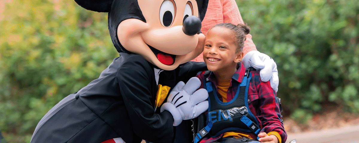 Une jeune fille en fauteuil roulant est accompagnée d'une personne vêtue d'une souris, soulignant ainsi l'esprit d'inclusion de Disneyland Paris et le soutien précieux apporté par un Accompagnant Handicap Disneyland Paris.