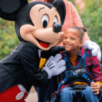 Une jeune fille en fauteuil roulant est accompagnée d'une personne vêtue d'une souris, soulignant ainsi l'esprit d'inclusion de Disneyland Paris et le soutien précieux apporté par un Accompagnant Handicap Disneyland Paris.