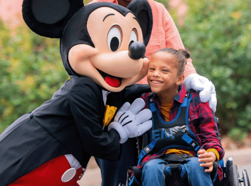 Une jeune fille en fauteuil roulant est accompagnée d'une personne vêtue d'une souris, soulignant ainsi l'esprit d'inclusion de Disneyland Paris et le soutien précieux apporté par un Accompagnant Handicap Disneyland Paris.