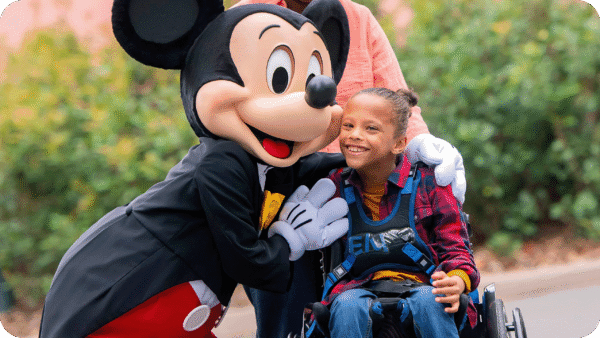 Une jeune fille en fauteuil roulant est accompagnée d'une personne vêtue d'une souris, soulignant ainsi l'esprit d'inclusion de Disneyland Paris et le soutien précieux apporté par un Accompagnant Handicap Disneyland Paris.