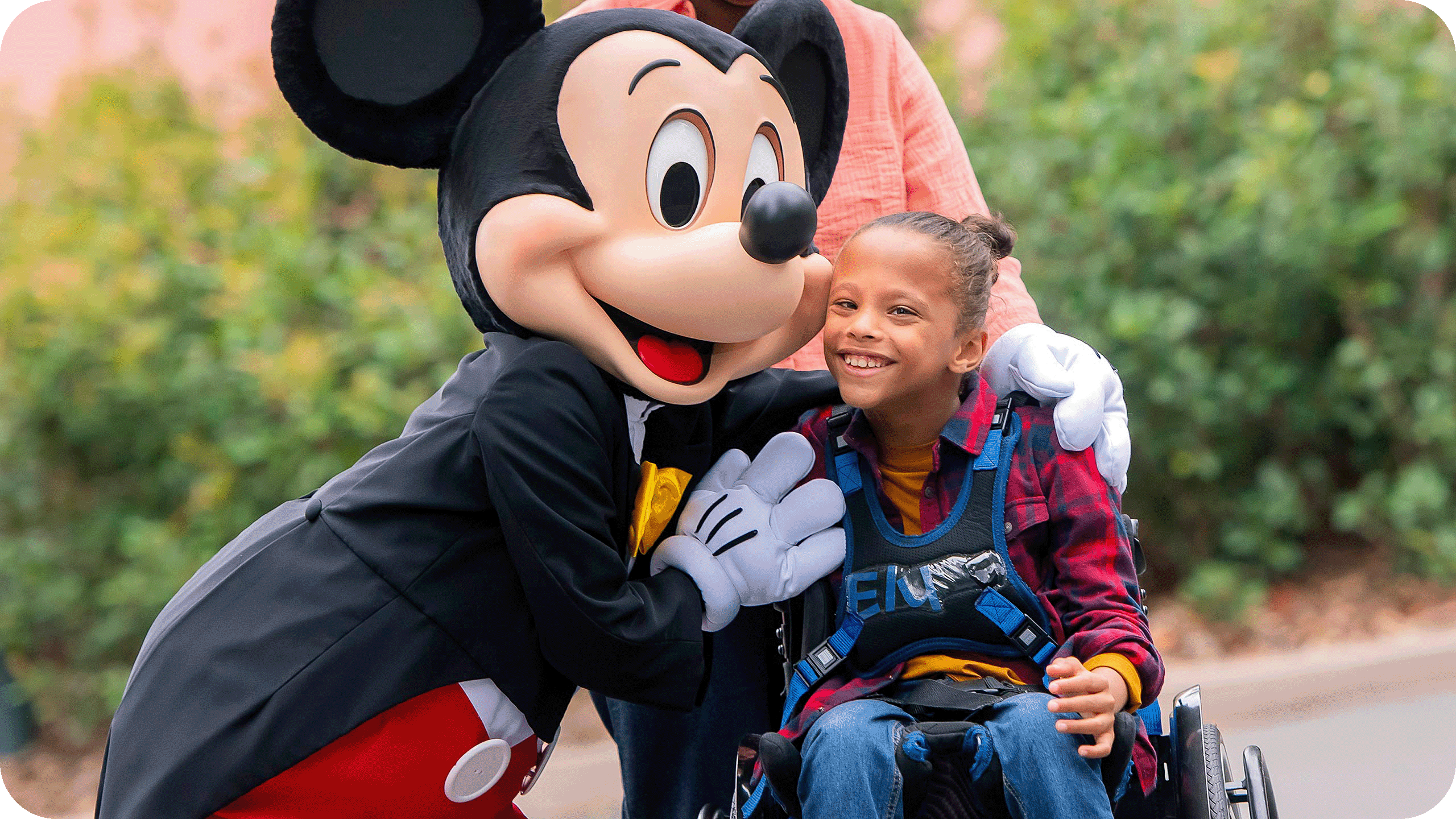 Une jeune fille en fauteuil roulant est accompagnée d'une personne vêtue d'une souris, soulignant ainsi l'esprit d'inclusion de Disneyland Paris et le soutien précieux apporté par un Accompagnant Handicap Disneyland Paris.
