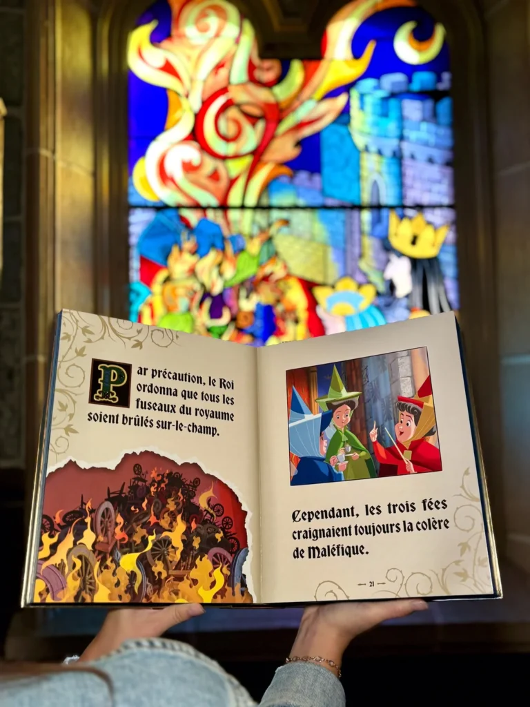 Une personne ouvre l'Édition Limitée du Grimoire De La Belle Au Bois Dormant, dont les illustrations féeriques brillent sous un vitrail coloré en arrière-plan.