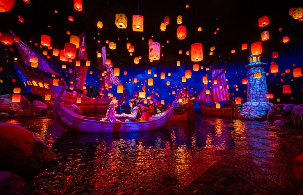 Un couple est assis dans un bateau sur un lac à Tokyo Disneyland, entouré de lanternes flottantes, de bateaux illuminés et d'un phare sous un ciel étoilé dans l'une des attractions les plus populaires du parc.