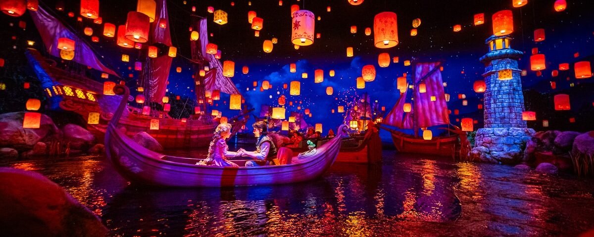 Un couple est assis dans un bateau sur un lac à Tokyo Disneyland, entouré de lanternes flottantes, de bateaux illuminés et d'un phare sous un ciel étoilé dans l'une des attractions les plus populaires du parc.