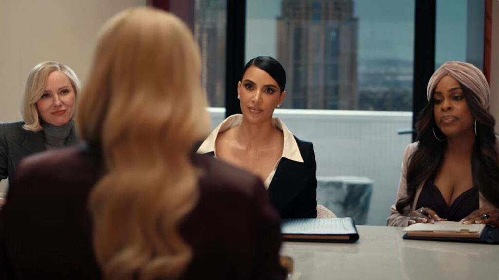 Trois femmes sont assises à une table de conférence et font face à une quatrième femme aux longs cheveux blonds, qui tourne le dos à la caméra, dans un bureau moderne où l'on aperçoit des bâtiments urbains, comme dans une scène de All's Fair sur Disney+.