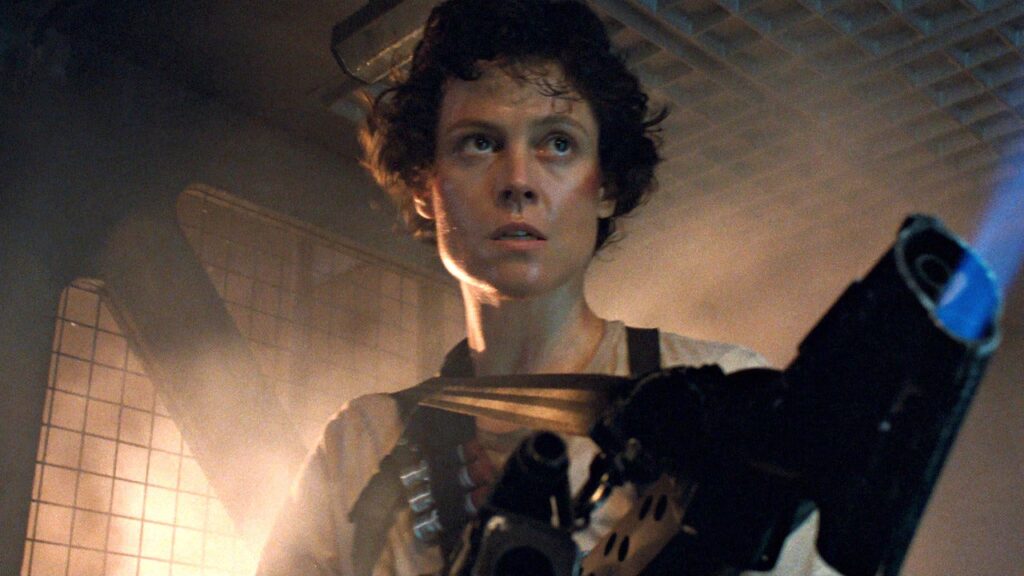 Une personne aux cheveux courts et bouclés se tient dans un environnement industriel faiblement éclairé, tenant une arme de grande taille, l'air alerte et concentré - rappelant la présence emblématique de Sigourney Weaver dans Alien.