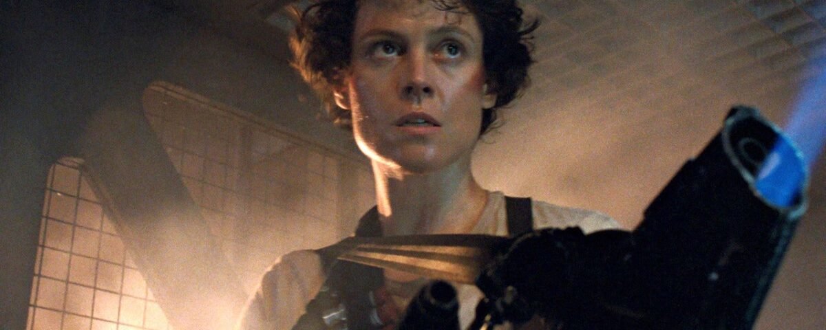 Une personne aux cheveux courts et bouclés se tient dans un environnement industriel faiblement éclairé, tenant une arme de grande taille, l'air alerte et concentré - rappelant la présence emblématique de Sigourney Weaver dans Alien.