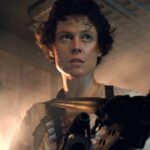 Une personne aux cheveux courts et bouclés se tient dans un environnement industriel faiblement éclairé, tenant une arme de grande taille, l'air alerte et concentré - rappelant la présence emblématique de Sigourney Weaver dans Alien.