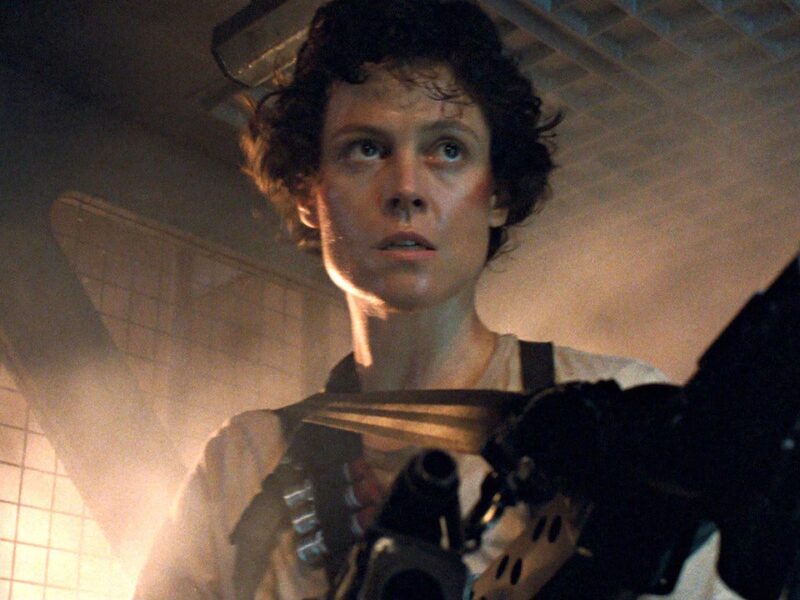 Une personne aux cheveux courts et bouclés se tient dans un environnement industriel faiblement éclairé, tenant une arme de grande taille, l'air alerte et concentré - rappelant la présence emblématique de Sigourney Weaver dans Alien.