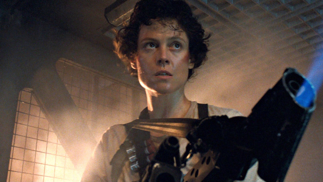 Une personne aux cheveux courts et bouclés se tient dans un environnement industriel faiblement éclairé, tenant une arme de grande taille, l'air alerte et concentré - rappelant la présence emblématique de Sigourney Weaver dans Alien.