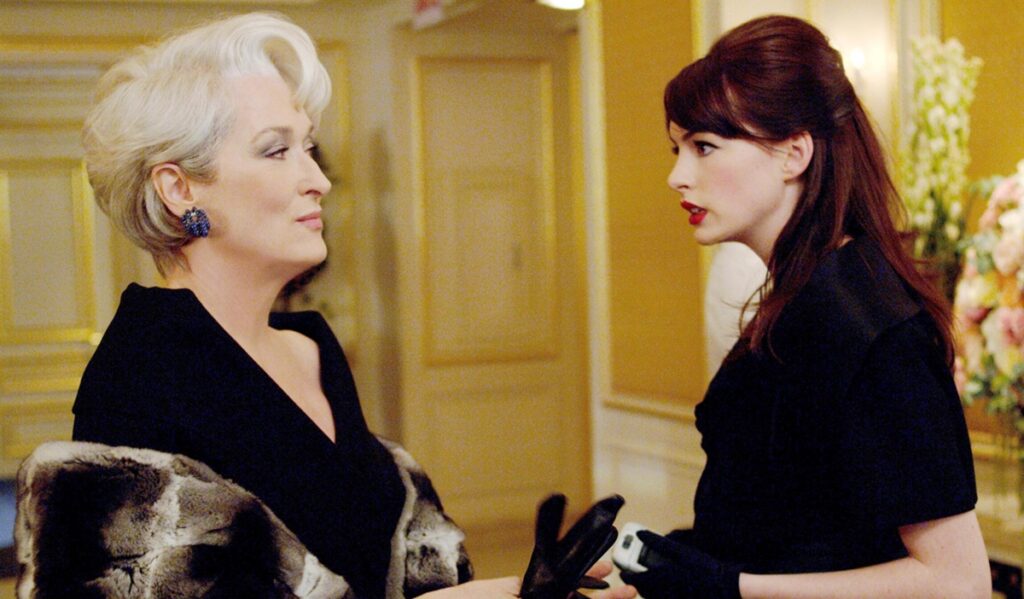 Deux femmes se font face dans un cadre formel ; l'une, qui rappelle Meryl Streep dans Le Diable S'habille En Prada, porte un manteau de fourrure et des gants, tandis que l'autre, qui reprend le style de Lady Gaga, tient un téléphone et porte des vêtements sombres.