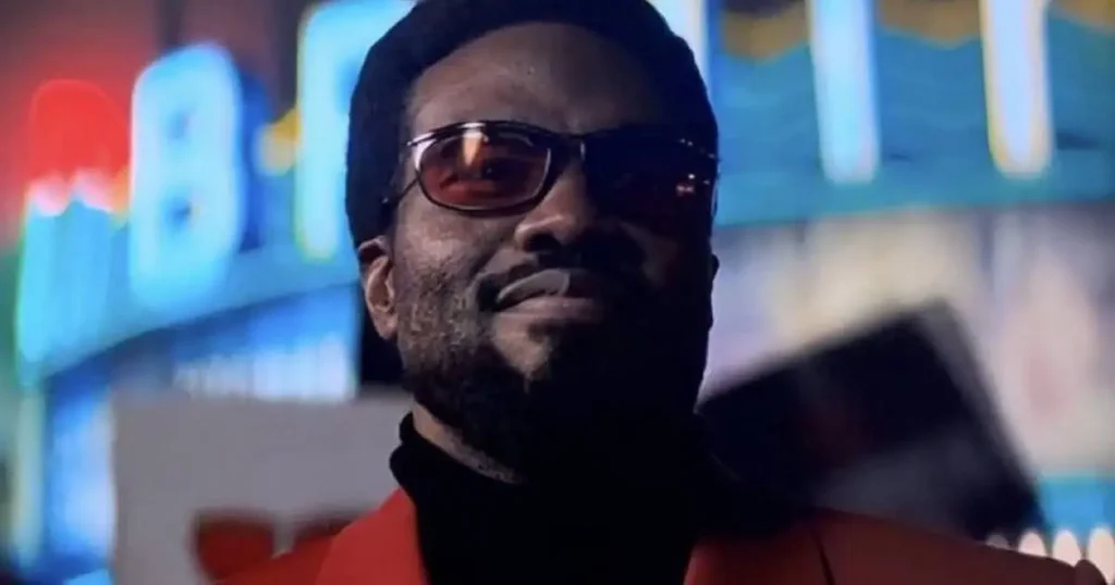 Un homme portant des lunettes de soleil et une veste rouge se tient devant des néons colorés et flous la nuit, dégageant l'assurance froide de Wonder Man de la série Disney+ de Marvel.