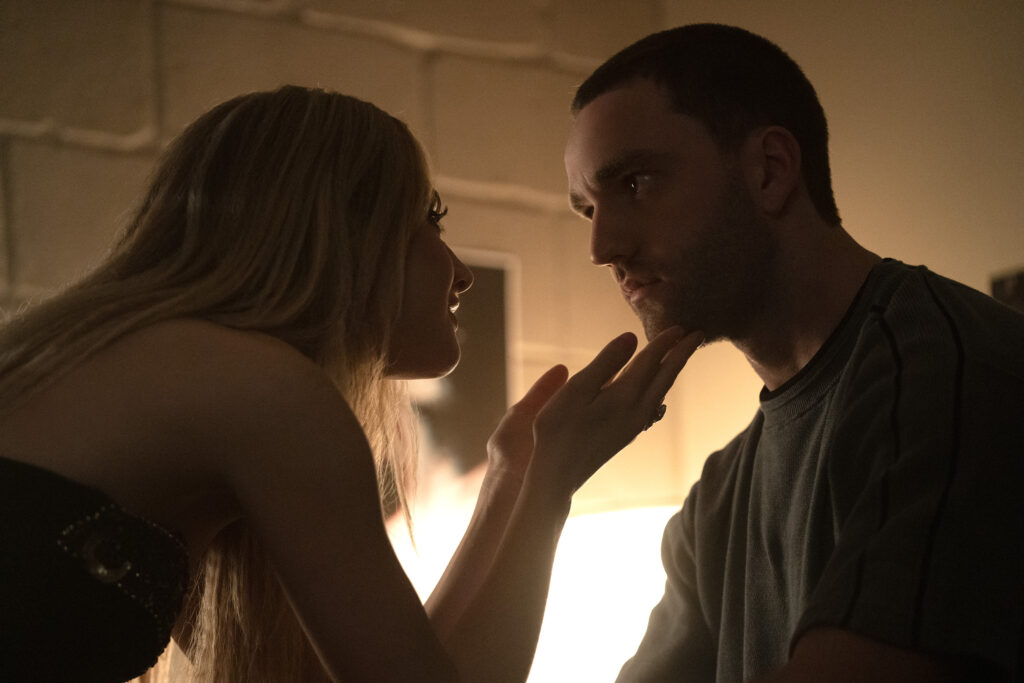 Une femme aux cheveux longs touche délicatement le menton d'un homme assis, tous deux se faisant face dans une pièce faiblement éclairée, faisant écho à un moment intime de Tell Me Lies S3 sur Disney+.