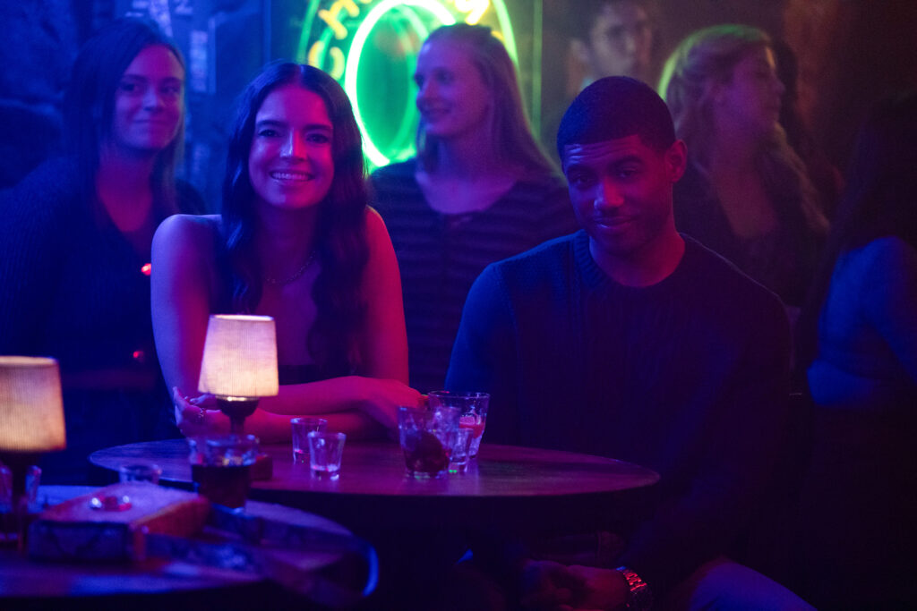 Deux personnes sont assises à une table ronde dans un bar faiblement éclairé, avec des boissons et des petites lampes sur la table. D'autres clients et une enseigne au néon sont visibles en arrière-plan, ce qui est parfait pour une scène de casting de Tell Me Lies S3 pour Disney+.