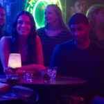 Deux personnes sont assises à une table ronde dans un bar faiblement éclairé, avec des boissons et des petites lampes sur la table. D'autres clients et une enseigne au néon sont visibles en arrière-plan, ce qui est parfait pour une scène de casting de Tell Me Lies S3 pour Disney+.