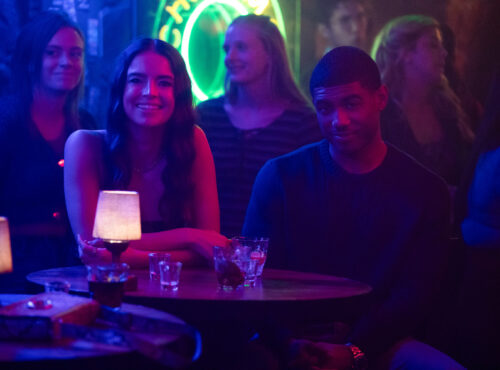 Deux personnes sont assises à une table ronde dans un bar faiblement éclairé, avec des boissons et des petites lampes sur la table. D'autres clients et une enseigne au néon sont visibles en arrière-plan, ce qui est parfait pour une scène de casting de Tell Me Lies S3 pour Disney+.