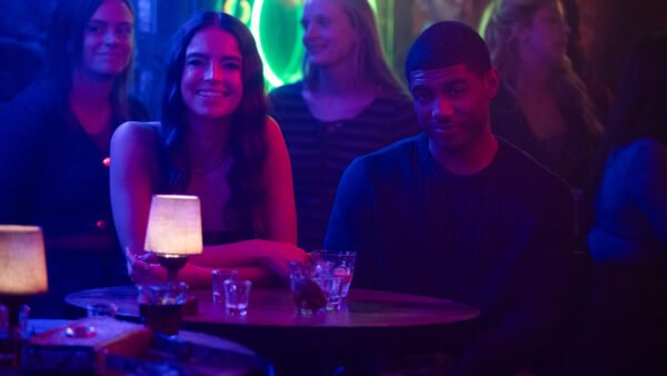 Deux personnes sont assises à une table ronde dans un bar faiblement éclairé, avec des boissons et des petites lampes sur la table. D'autres clients et une enseigne au néon sont visibles en arrière-plan, ce qui est parfait pour une scène de casting de Tell Me Lies S3 pour Disney+.