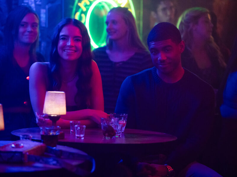 Deux personnes sont assises à une table ronde dans un bar faiblement éclairé, avec des boissons et des petites lampes sur la table. D'autres clients et une enseigne au néon sont visibles en arrière-plan, ce qui est parfait pour une scène de casting de Tell Me Lies S3 pour Disney+.