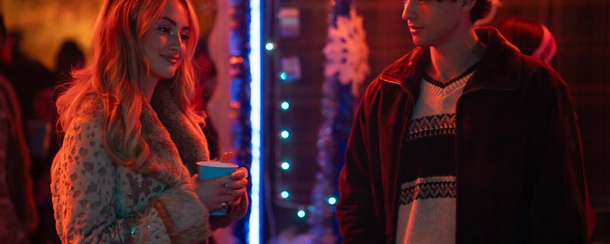 Deux personnes discutent lors d'une fête peu éclairée, décorée de guirlandes, de flocons de neige et de lumières bleues. Vêtue de vêtements d'hiver, la femme tient une tasse. Cette scène ressemble au casting de Tell Me Lies S3, prêt pour ses débuts sur Disney+.