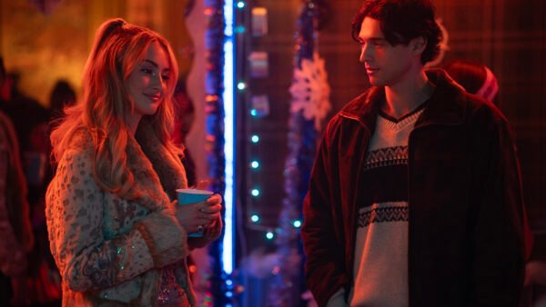 Deux personnes discutent lors d'une fête peu éclairée, décorée de guirlandes, de flocons de neige et de lumières bleues. Vêtue de vêtements d'hiver, la femme tient une tasse. Cette scène ressemble au casting de Tell Me Lies S3, prêt pour ses débuts sur Disney+.