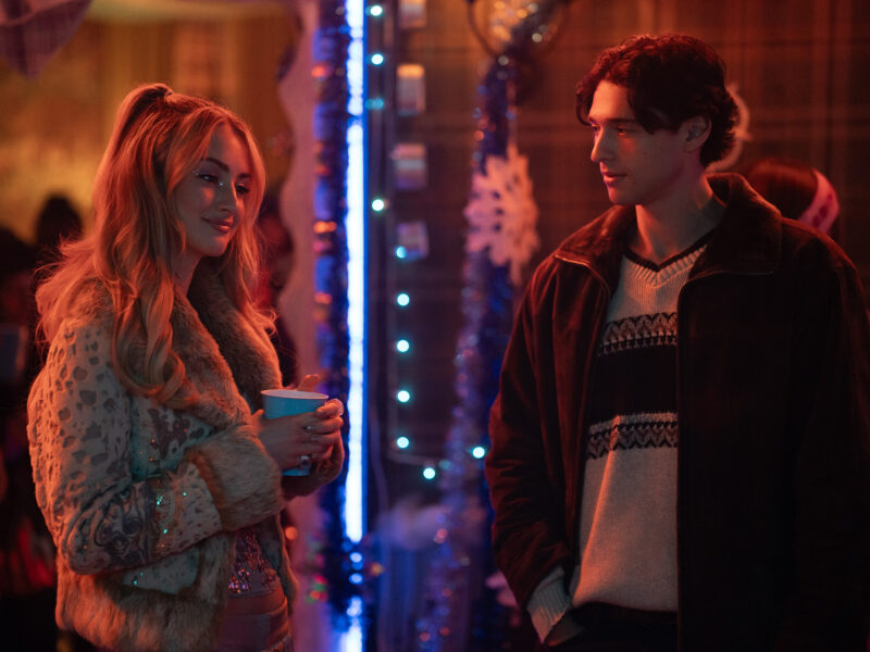 Deux personnes discutent lors d'une fête peu éclairée, décorée de guirlandes, de flocons de neige et de lumières bleues. Vêtue de vêtements d'hiver, la femme tient une tasse. Cette scène ressemble au casting de Tell Me Lies S3, prêt pour ses débuts sur Disney+.