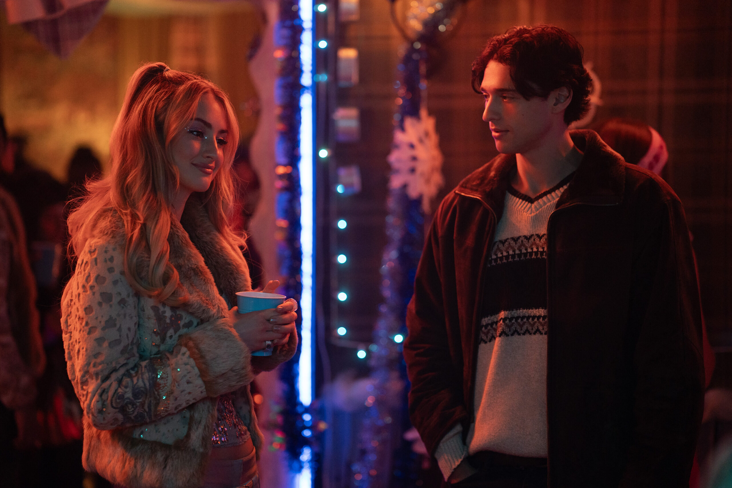 Deux personnes discutent lors d'une fête peu éclairée, décorée de guirlandes, de flocons de neige et de lumières bleues. Vêtue de vêtements d'hiver, la femme tient une tasse. Cette scène ressemble au casting de Tell Me Lies S3, prêt pour ses débuts sur Disney+.