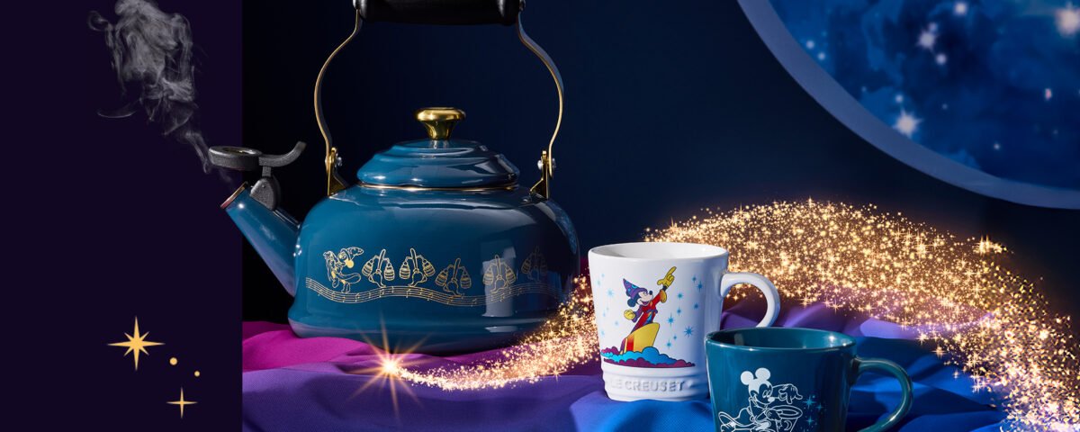 Une bouilloire Le Creuset bleue avec des symboles de Fantasia et deux tasses Disney sont posées sur un tissu coloré. Les ustensiles Disney brillent tandis que des paillettes dorées tourbillonnent autour d'eux sur un fond sombre.
