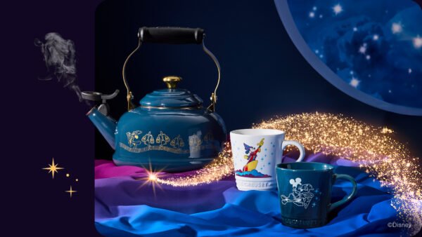 Une bouilloire Le Creuset bleue avec des symboles de Fantasia et deux tasses Disney sont posées sur un tissu coloré. Les ustensiles Disney brillent tandis que des paillettes dorées tourbillonnent autour d'eux sur un fond sombre.