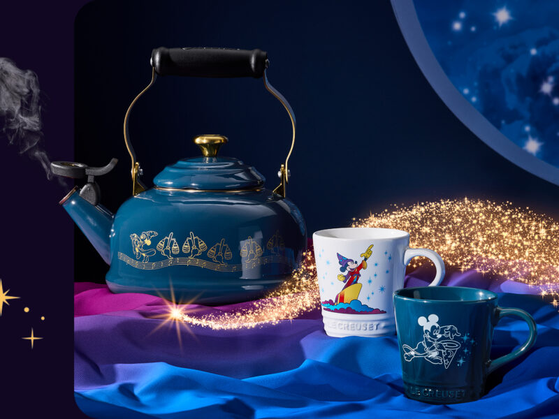 Une bouilloire Le Creuset bleue avec des symboles de Fantasia et deux tasses Disney sont posées sur un tissu coloré. Les ustensiles Disney brillent tandis que des paillettes dorées tourbillonnent autour d'eux sur un fond sombre.
