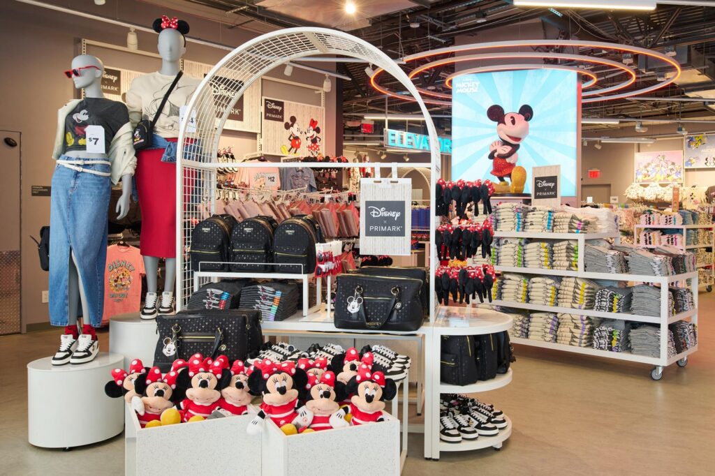 Magasin de détail Primark sur le thème de Disney, présentant des peluches Minnie Mouse, des sacs à dos, des t-shirts pliés, des chaussures et des mannequins vêtus de vêtements Disney, le tout dans un cadre abordable. Grande image de Mickey Mouse en arrière-plan.