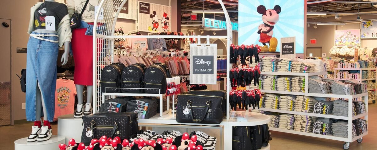 Magasin de détail Primark sur le thème de Disney, présentant des peluches Minnie Mouse, des sacs à dos, des t-shirts pliés, des chaussures et des mannequins vêtus de vêtements Disney, le tout dans un cadre abordable. Grande image de Mickey Mouse en arrière-plan.