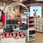 Magasin de détail Primark sur le thème de Disney, présentant des peluches Minnie Mouse, des sacs à dos, des t-shirts pliés, des chaussures et des mannequins vêtus de vêtements Disney, le tout dans un cadre abordable. Grande image de Mickey Mouse en arrière-plan.