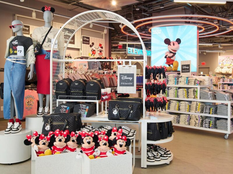 Magasin de détail Primark sur le thème de Disney, présentant des peluches Minnie Mouse, des sacs à dos, des t-shirts pliés, des chaussures et des mannequins vêtus de vêtements Disney, le tout dans un cadre abordable. Grande image de Mickey Mouse en arrière-plan.