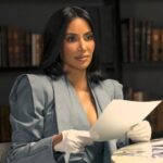 Une femme aux cheveux bruns, vêtue d'un blazer et de gants blancs, rappelant le style soigné de Kim Kardashian, tient une feuille de papier, assise à une table avec un verre, devant des étagères.