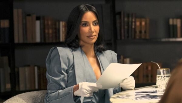 Une femme aux cheveux bruns, vêtue d'un blazer et de gants blancs, rappelant le style soigné de Kim Kardashian, tient une feuille de papier, assise à une table avec un verre, devant des étagères.