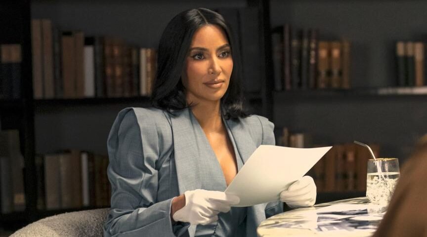 Une femme aux cheveux bruns, vêtue d'un blazer et de gants blancs, rappelant le style soigné de Kim Kardashian, tient une feuille de papier, assise à une table avec un verre, devant des étagères.