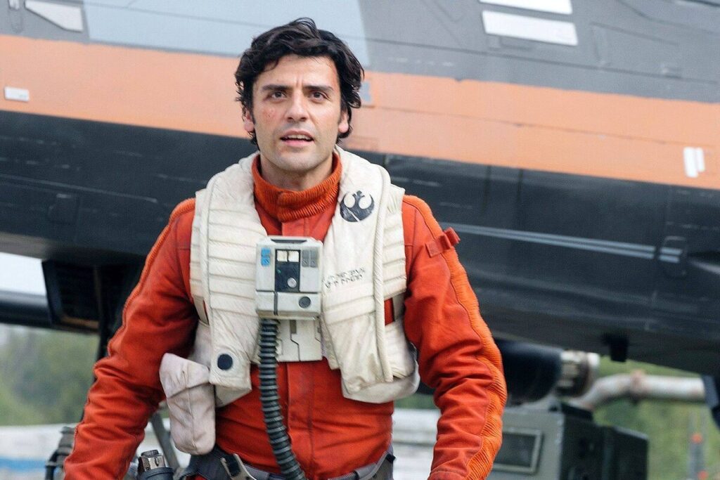 Un homme vêtu d'une combinaison de vol et d'un gilet orange se tient devant un vaisseau spatial, un appareil de communication autour du cou - rappelant le rôle emblématique d'Oscar Isaac dans La Guerre des étoiles.