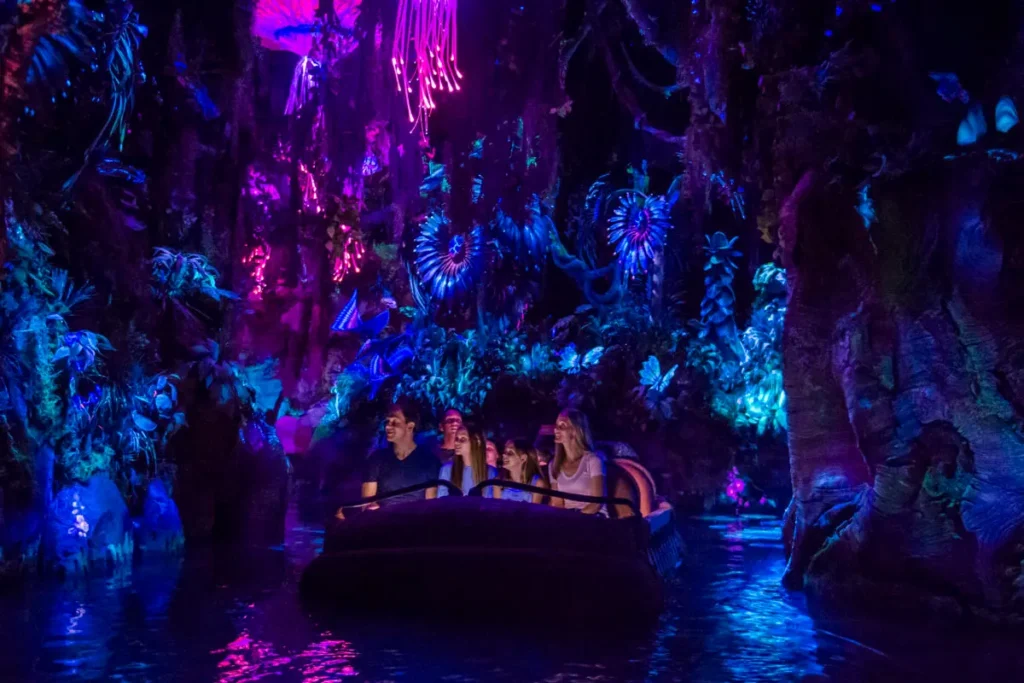Un groupe de personnes traverse en bateau une forêt bioluminescente aux éclairages violets, bleus et roses dans le monde enchanteur d'Avatar Land à Disneyland Resort.