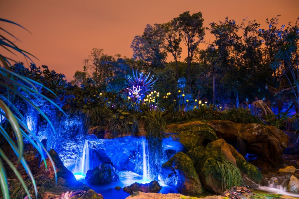 Scène nocturne d'un paysage bioluminescent artificiel avec des plantes, des cascades et des arbres lumineux sous un ciel orange - comme si vous exploriez Avatar Land, la nouvelle attraction immersive de Disneyland Resort.