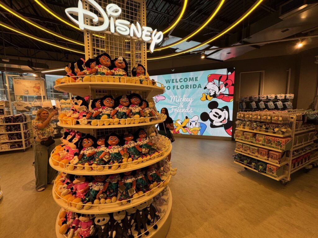 Un présentoir de peluches Disney est disposé sur un stand circulaire à l'intérieur de Primark, avec un panneau "Welcome to Florida" et des personnages Disney sur un écran en arrière-plan, mettant en valeur le mode abordable pour tous les fans.