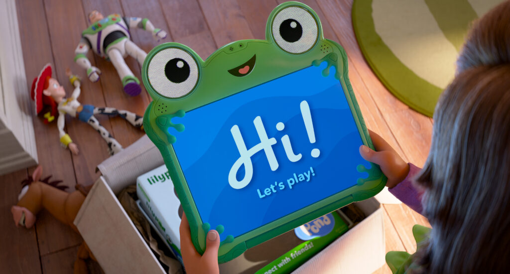 Un enfant tient une tablette verte en forme de grenouille affichant le message "Hi ! Let's play !" devant une boîte contenant des jouets et des livres sur un sol en bois, faisant allusion à la magie de Pixar et à l'excitation pour Toy Story 5.