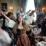 Un groupe de personnes en tenue historique se tient dans une pièce, beaucoup avec les bras levés et des visages expressifs, tandis qu'une femme au centre - évoquant Amanda Seyfried dans un biopic - regarde vers le haut. Inspiré par Le Testament D'Ann Lee.