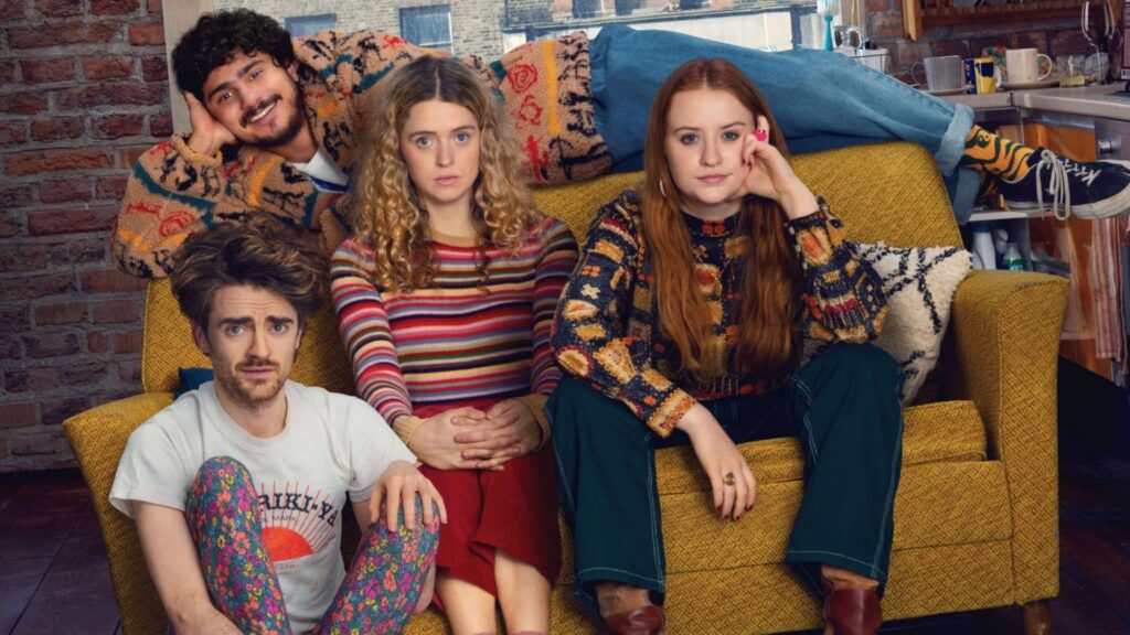 Quatre jeunes adultes sont assis et adossés à un canapé jaune dans un salon, portant des vêtements colorés et regardant la caméra avec des expressions neutres, comme s'ils posaient pour une série comique telle que Extraordinary de Disney.