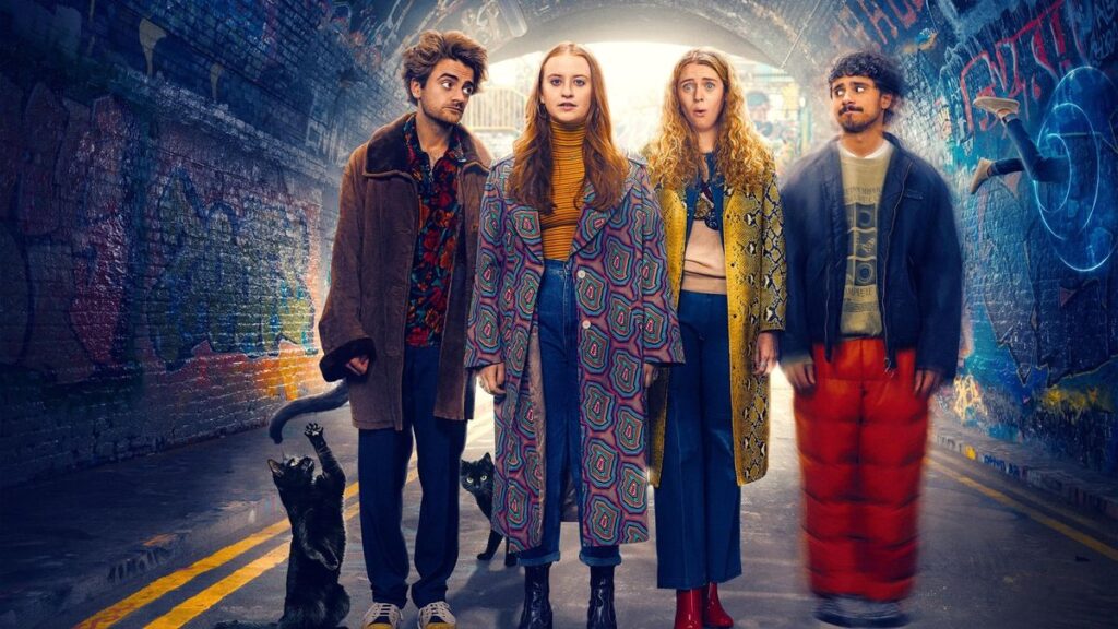 Quatre jeunes adultes se tiennent dans un tunnel couvert de graffitis, deux chats noirs à leurs pieds, tous vêtus de tenues colorées et excentriques, regardant vers l'avant avec des expressions neutres, comme s'ils étaient prêts à vivre une aventure extraordinaire dans une série comique de Disney.