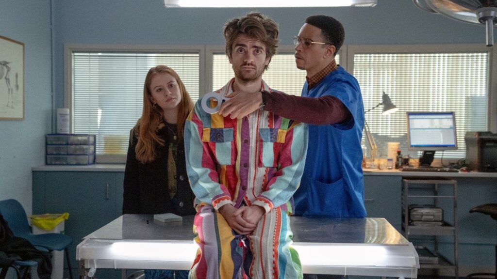 Un homme vêtu d'une tenue à rayures colorées est assis sur une table d'hôpital tandis qu'un médecin, tout droit sorti d'une série comique de Disney, tient du ruban adhésif médical près de son épaule ; une femme extraordinaire se tient à l'arrière-plan et observe.