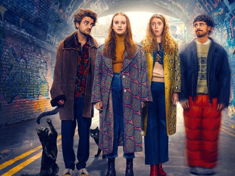 Quatre jeunes adultes se tiennent dans un tunnel couvert de graffitis, deux chats noirs à leurs pieds, tous vêtus de tenues colorées et excentriques, regardant vers l'avant avec des expressions neutres, comme s'ils étaient prêts à vivre une aventure extraordinaire dans une série comique de Disney.