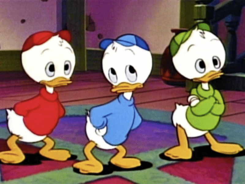 Trois canetons de dessin animé, dont Riri, portent des chemises et des casquettes rouges, bleues et vertes. Ils se tiennent à l'intérieur, les mains derrière le dos, face à l'avant.