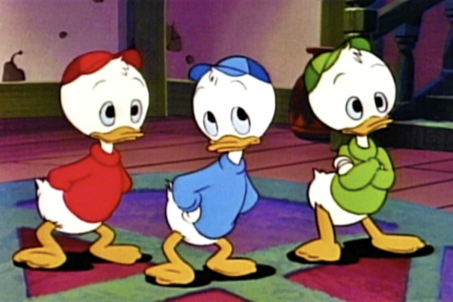 Trois canetons de dessin animé, dont Riri, portent des chemises et des casquettes rouges, bleues et vertes. Ils se tiennent à l'intérieur, les mains derrière le dos, face à l'avant.