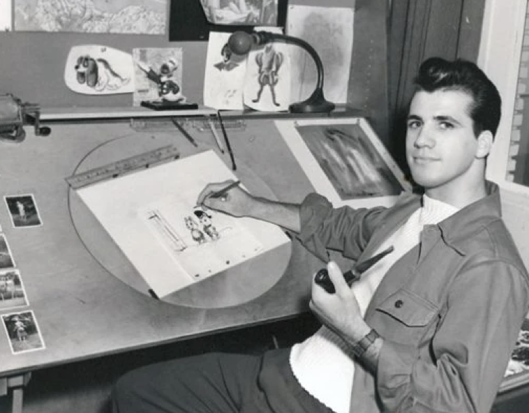 Un homme est assis à un bureau de dessin, tenant une pipe et esquissant une caricature sur papier, avec des œuvres d'art et des outils disposés autour de lui - une scène qui rappelle celle d'un artiste se préparant à la première de Robin des Bois, le 8 novembre 1973.